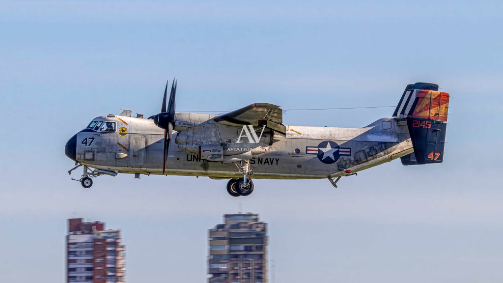 Un avión militar de la Marina de los Estados Unidos volando sobre una ciudad, con un diseño de camuflaje y número visible en la parte delantera.